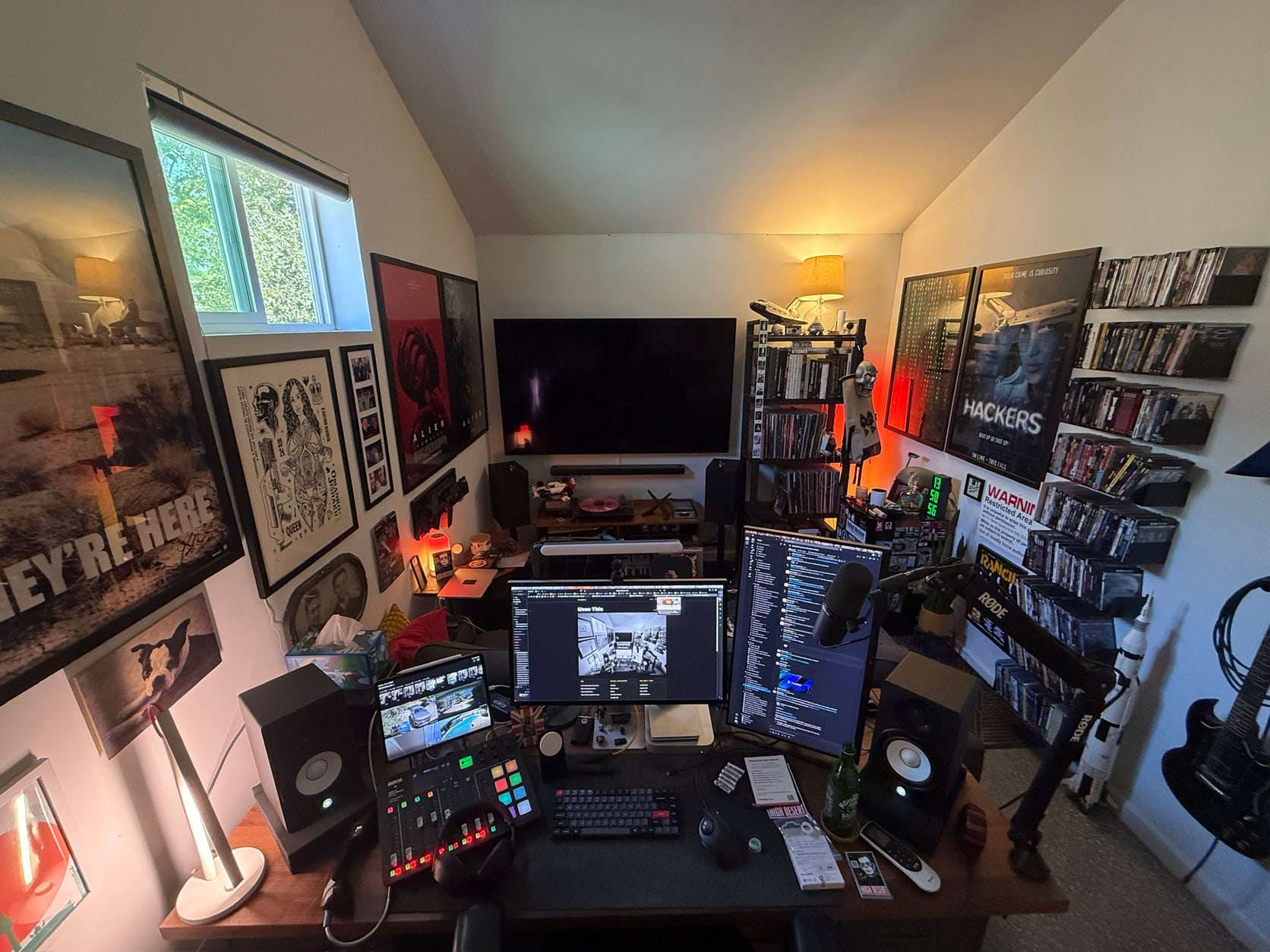 Wide-angle view of a home workspace with multiple displays, audio gear, a television, framed posters, and shelves of physical media.